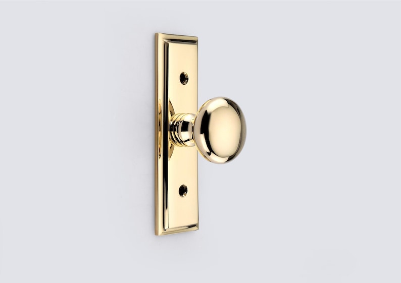 May include: A polished brass door knob set featuring a round, spherical knob attached to a rectangular backplate with beveled edges. The backplate includes two circular screw holes positioned vertically above and below the central knob mechanism.
