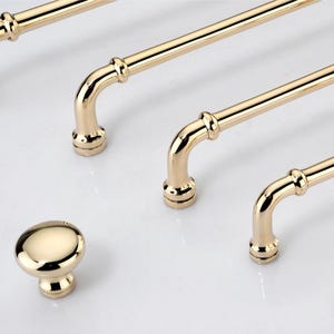 May include: A collection of polished gold-tone cabinet hardware including four long, tubular bar pulls with rounded ends and a single matching round cabinet knob. Each piece features a smooth, reflective metallic finish and circular base mounts.