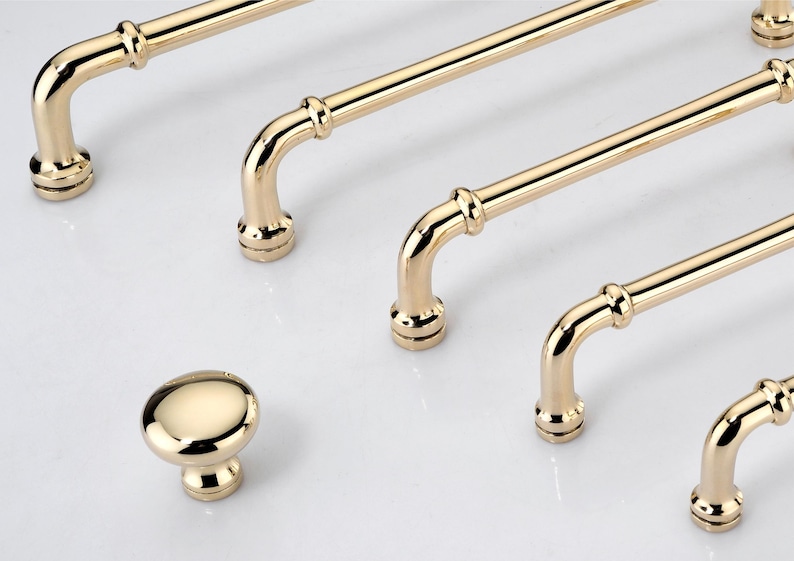 May include: A collection of polished gold-tone cabinet hardware including four long, tubular bar pulls with rounded ends and a single matching round cabinet knob. Each piece features a smooth, reflective metallic finish and circular base mounts.