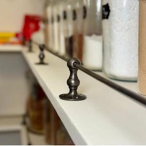 Könnte beinhalten: Eine dunkelbronzefarbene Metall-Regalschiene mit dekorativen Halterungen. Die Schiene ist auf einem weißen Regal montiert, im Hintergrund befinden sich Gläser mit Lebensmitteln. Die Regalschiene soll verhindern, dass Gegenstände herunterfallen.