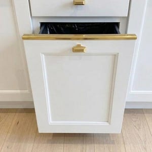 May include: A white kitchen cabinet drawer with a gold-toned metal handle and a gold-toned trim along the top edge. The drawer is partially pulled open, revealing a black plastic trash bin liner inside the cabinet compartment.