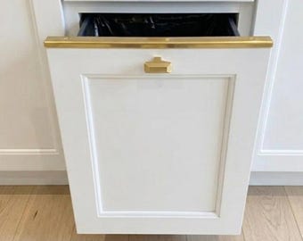 May include: A white kitchen cabinet drawer with a gold-toned metal handle and a gold-toned trim along the top edge. The drawer is partially pulled open, revealing a black plastic trash bin liner inside the cabinet compartment.