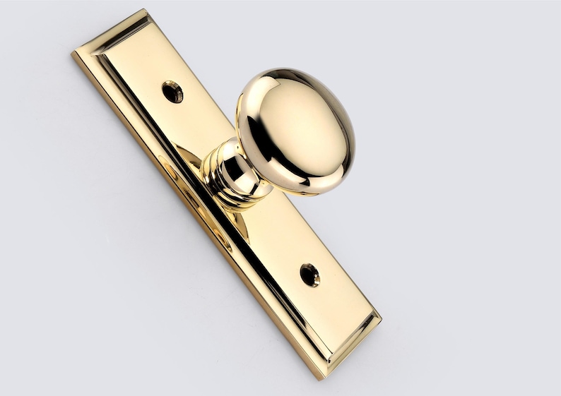 May include: A polished brass door knob set featuring a round, smooth knob attached to a rectangular backplate with beveled edges. The backplate includes two circular screw holes, one positioned above and one below the central knob assembly.