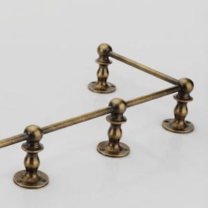 May include: A wall-mounted rail system featuring a metallic antique brass finish. The hardware consists of three vertical supports with circular base plates and a horizontal rod that extends across the top, secured by spherical finial connectors.