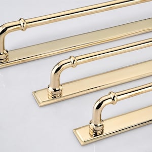 May include: A set of polished gold-toned metal cabinet pulls, each featuring a long, flat rectangular backplate and a rounded, tubular handle attached with two curved supports. The hardware has a reflective, smooth finish and a clean design.
