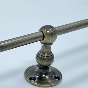 May include: A metal towel bar with an antique brass finish featuring a horizontal rod supported by a central post with a spherical accent and a circular mounting base with three visible screw holes for secure attachment to a wall surface.