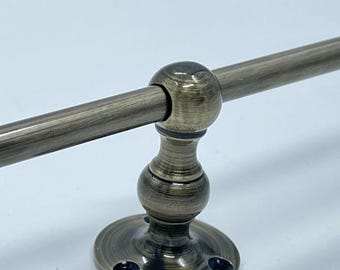 May include: A metal towel bar with an antique brass finish featuring a horizontal rod supported by a central post with a spherical accent and a circular mounting base with three visible screw holes for secure attachment to a wall surface.