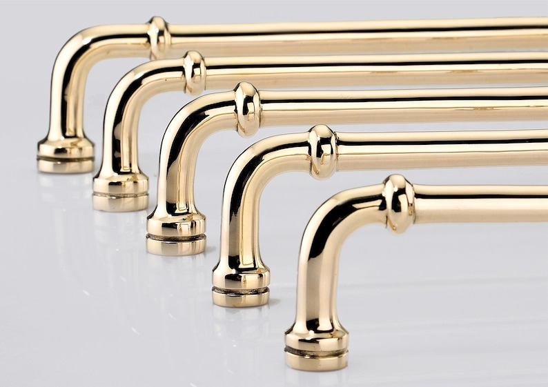 May include: A series of five polished gold-toned metal cabinet pulls arranged in a row. Each handle features a smooth, curved bar design with a rounded post at each end, finished with a decorative ring detail near the base of the mounting points.