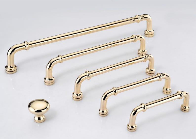 May include: A collection of polished gold-toned cabinet hardware, including one round knob and five arched bar pulls of varying lengths. Each piece features a smooth, reflective metallic finish with cylindrical bars and circular mounting bases.