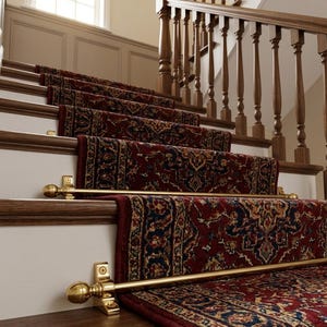 May include: A staircase featuring a dark red patterned runner rug secured by polished brass stair rods with spherical finials. The wooden steps have white risers and a dark wood handrail with vertical balusters extending up the staircase.