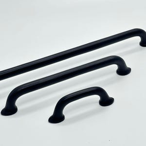 May include: Three black cabinet pulls of different sizes against a white backdrop. The pulls have a curved design with rounded ends. The largest is a long bar, the others are shorter, curved handles. Measurements are in cm.