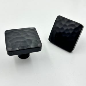 Puede incluir: Dos tiradores cuadrados negros para armarios con un aspecto texturizado y martillado. Los tiradores tienen un acabado oscuro y un diseño sencillo y moderno. Se colocan sobre un fondo blanco, resaltando su forma y textura. Adecuado para muebles o herrajes.