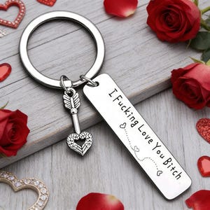 Puede incluir: Llavero plateado con una etiqueta rectangular que dice "I Fucking Love You Bitch". El llavero tiene un dije de flecha en forma de corazón y un anillo grande. Rosas rojas y decoraciones en forma de corazón rodean el llavero.