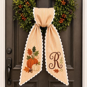 May include: A beige and white striped fabric door hanger with scalloped edges. The left side features an embroidered orange pumpkin, a white pumpkin, and autumn leaves. The right side displays a large brown embroidered letter R with leaves.