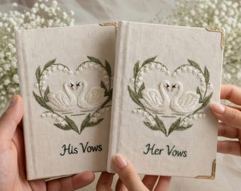 May include: Two off-white fabric-covered vow books with gold-tone metal corners. Each features an embroidered heart-shaped wreath of green leaves and white flowers surrounding two white swans. One book reads His Vows, the other Her Vows.