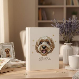 May include: A cream-coloured book featuring a detailed illustration of a dog's face, the name "Bubba" below, and gold stars. A framed photo of the dog is nearby. A vase of lavender and a cup are also on the table.