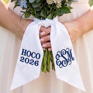 Könnte beinhalten: Ein weißes Satinband, das um einen Blumenstrauß gebunden ist. Das Band trägt den Text "HOCO 2026" und ein marineblaues Monogramm. Der Strauß enthält Grün und weiße und blaue Blumen.