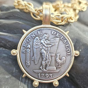 Puede incluir: Colgante dorado con un diseño detallado de moneda de plata. La moneda presenta una figura alada, un gallo y el año 1793. El colgante está sujeto a una cadena dorada. La moneda está engastada en un marco dorado con detalles decorativos.