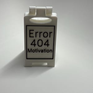 Könnte beinhalten: Ein kleines, weißes, rechteckiges Schild mit dem Text "Error 404 Motivation" in Schwarz. Das Schild hat einen Griff oben und einen Sockel zum Stehen. Das Schild befindet sich auf einer weißen Oberfläche.
