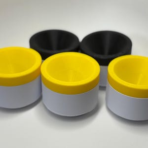 Op de afbeelding: Een verzameling kleine, cilindervormige containers met witte lichamen en felgele deksels. Er zijn ook twee zwarte, komvormige objecten aanwezig. De containers zijn gerangschikt op een wit oppervlak.