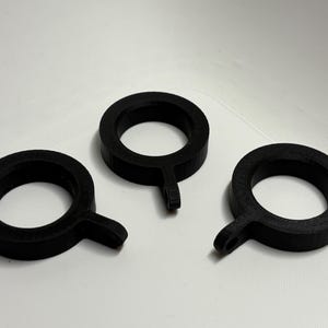 Könnte beinhalten: Drei schwarze, kreisförmige Gardinenringe mit einem kleinen, rechteckigen Ansatz an der Unterseite. Die Ringe bestehen aus einem matten Material und sind auf weißem Hintergrund angeordnet.