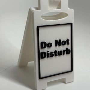 Könnte beinhalten: Ein weißes Kunststoffschild mit den schwarzen Worten "Do Not Disturb". Das Schild hat einen schwarzen Rahmen und einen Griff oben. Das Schild ist so konzipiert, dass es selbstständig steht. Das Schild ist aus Kunststoff.