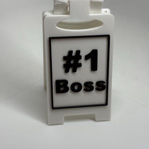 Może przedstawiać: Biały, składany znak z napisem "#1 Boss" w kolorze czarnym. Znak ma czarną obwódkę i uchwyt u góry. Zaprojektowany do stania, idealny na biurko.