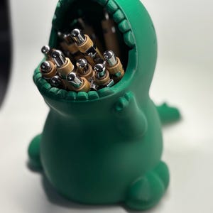 Op de afbeelding: Een groene bureau-organizer in de vorm van een dinosaurus, met een open mond vol pennen en potloden. De organizer is gemaakt van een effen groen materiaal en heeft een gladde afwerking. De pennen en potloden hebben zilveren en bamboe accenten.