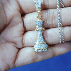 May include: A gold and silver pendant necklace featuring a chess king piece design. The pendant is encrusted with small, sparkling stones and has gold accents. The necklace chain is silver.