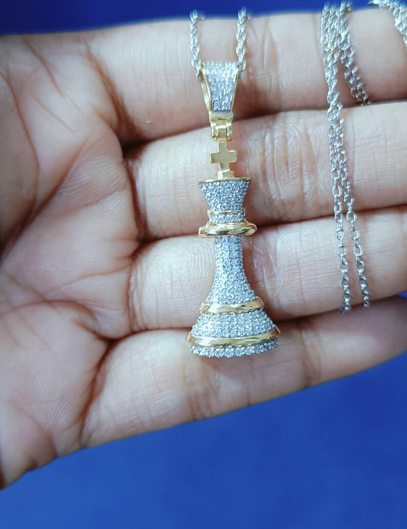 May include: A gold and silver pendant necklace featuring a chess king piece design. The pendant is encrusted with small, sparkling stones and has gold accents. The necklace chain is silver.