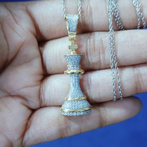 May include: A silver necklace with a pendant shaped like a chess king piece. The pendant is encrusted with small, clear stones and has gold-colored accents. The chain is also silver.