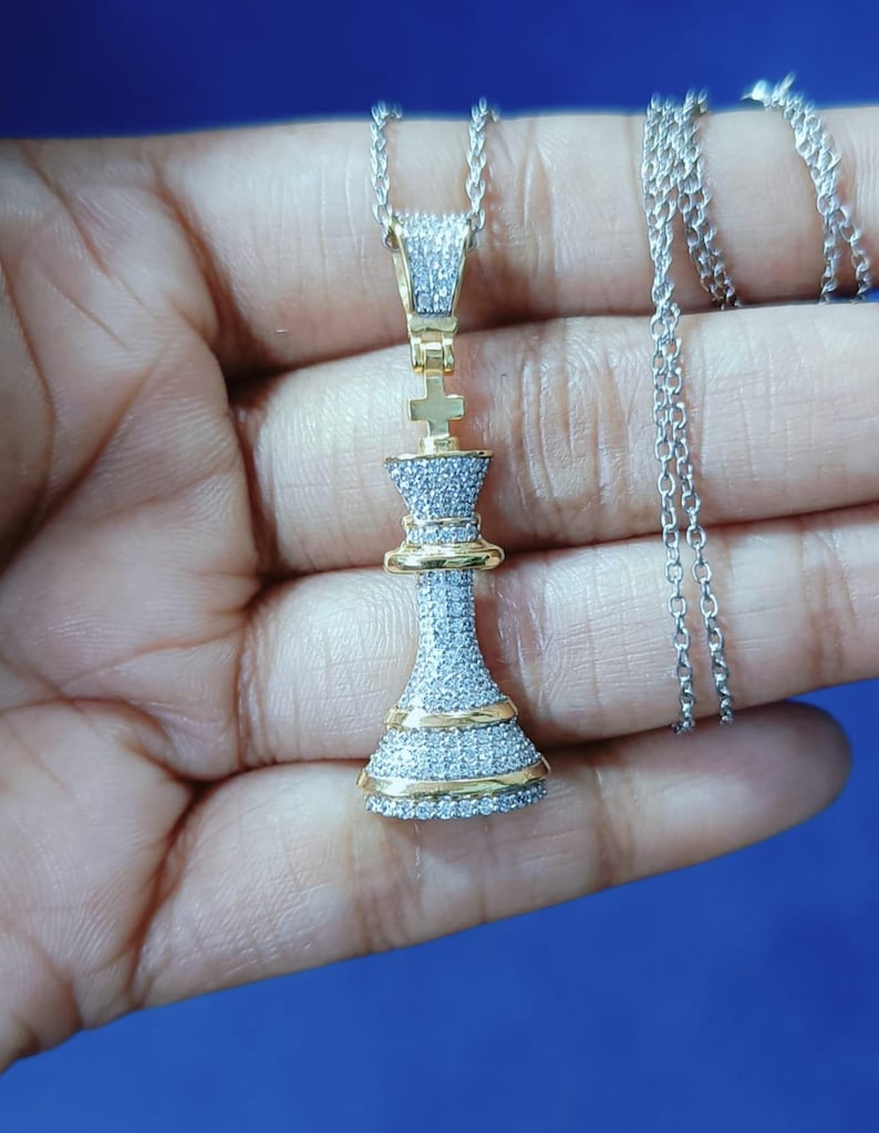 May include: A silver necklace with a pendant shaped like a chess king piece. The pendant is encrusted with small, clear stones and has gold-colored accents. The chain is also silver.