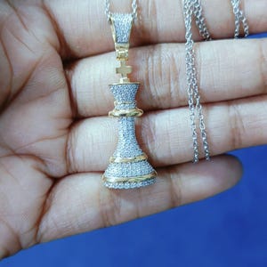 May include: A silver chain necklace with a pendant shaped like a chess piece, the king. The pendant is encrusted with small, clear stones and has gold-colored accents. The necklace is displayed against a blue background.