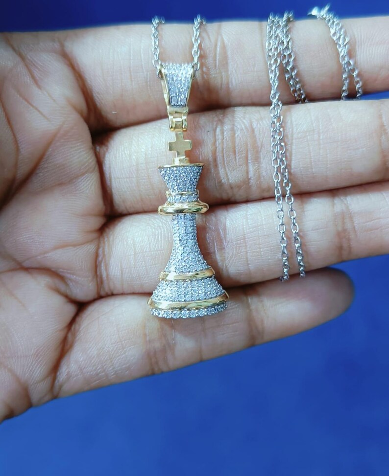 May include: A silver chain necklace with a pendant shaped like a chess piece, the king. The pendant is encrusted with small, clear stones and has gold-colored accents. The necklace is displayed against a blue background.