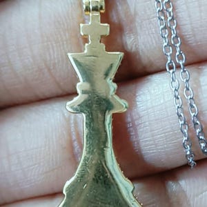 May include: Gold-colored pendant necklace featuring a chess king piece. The pendant is suspended from a silver-colored chain. The king piece is detailed with a cross on top.