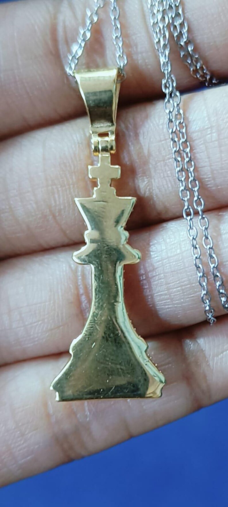 May include: Gold-colored pendant necklace featuring a chess king piece. The pendant is suspended from a silver-colored chain. The king piece is detailed with a cross on top.
