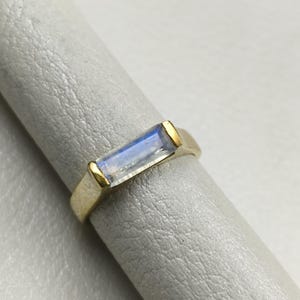 May include: A gold ring featuring a rectangular baguette-cut moonstone with a blue iridescent sheen. The gemstone is set horizontally in a smooth gold band, showcasing a clean, polished finish and a simple, elegant design.
