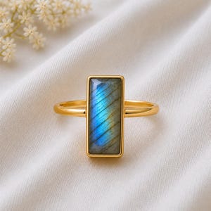 May include: A gold ring featuring a rectangular labradorite gemstone set vertically. The stone displays a prominent blue iridescent flash across its surface, framed by a smooth gold bezel. The band is thin, polished gold, creating a sleek appearance.