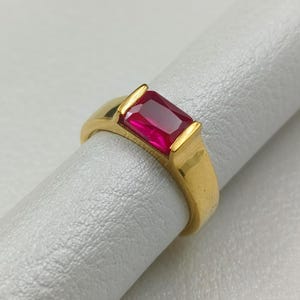 May include: A gold ring featuring a rectangular, vibrant red gemstone. The ring has a classic design with a thick band and a bezel setting that securely holds the gemstone. The ring is shown on a white surface.