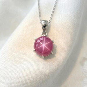 May include: A silver necklace featuring a round, pink gemstone pendant. The gemstone displays a star-shaped pattern and is secured by six silver prongs. The fine chain hangs against a white backdrop.