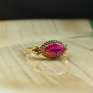 Puede incluir: Un anillo de oro con una piedra preciosa de color rubí con corte marquesa. El anillo presenta un diseño de hojas en los laterales y un borde negro con cuentas alrededor de la piedra preciosa. El anillo está sobre una superficie de madera clara.