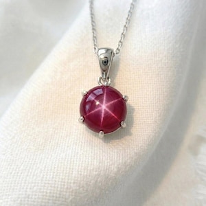 Puede incluir: Un collar de plata con un colgante de gema redonda de color rojo intenso. La gema presenta un patrón en forma de estrella y está sujeta por un engaste de plata con pequeñas garras. La cadena es delicada y plateada.