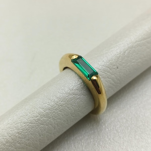May include: A gold ring with a rectangular emerald-cut green gemstone. The ring has a smooth, rounded band and is set with the emerald in a horizontal orientation. The ring is displayed on a light gray surface.