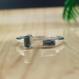 May include: A silver ring with two rectangular, dark blue gemstones. The ring has an open design, with the gemstones set on either side of the opening. The ring is displayed on a reflective surface.