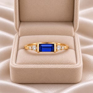 May include: A gold ring featuring a central rectangular baguette-cut blue sapphire flanked by two small round brilliant-cut diamonds on each side, resting inside a beige velvet jewelry box with a soft, satin-lined interior.
