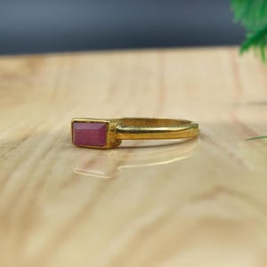 Pu&ograve; includere: Un anello d'oro con una pietra rettangolare color rubino. L'anello ha una fascia sottile e lucida, e la pietra &egrave; incastonata in una lunetta rettangolare. L'anello &egrave; esposto su una superficie di legno chiaro.