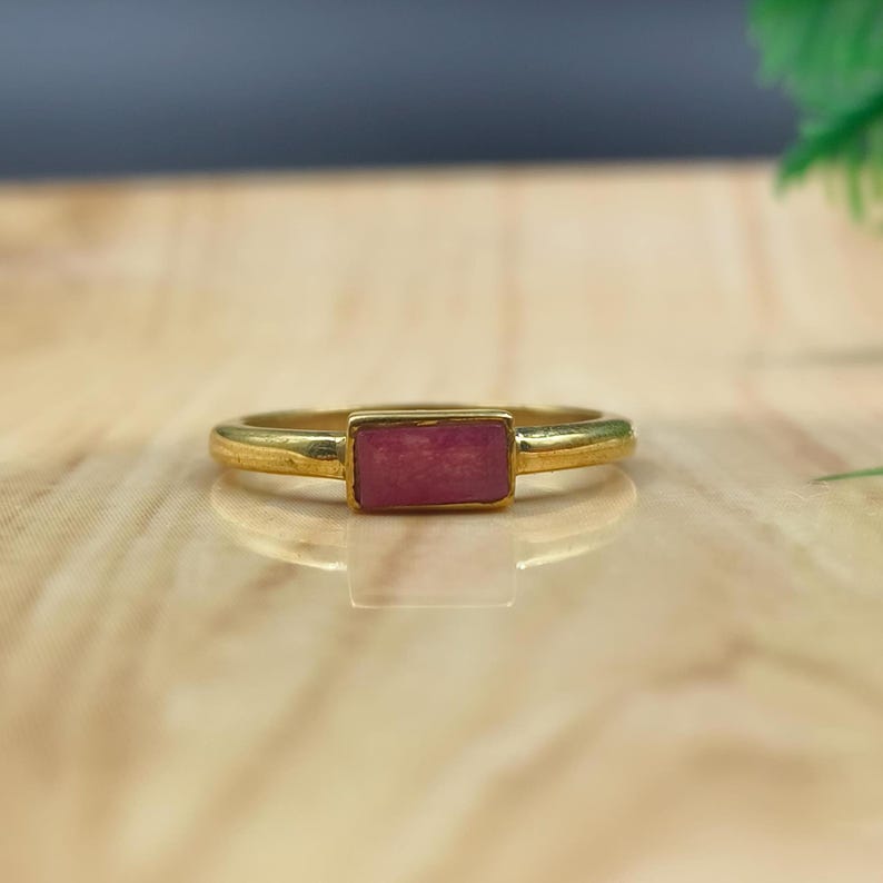 Pu&ograve; includere: Un anello color oro con una pietra rettangolare rosa-rossa. La fascia dell'anello &egrave; liscia e semplice, e la pietra &egrave; incastonata in un castone a lunetta. L'anello &egrave; probabilmente in metallo placcato oro. Questo anello &egrave; un gioiello.