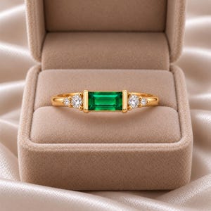 May include: A gold ring featuring a central rectangular baguette-cut green gemstone flanked by two round brilliant-cut clear stones on each side. The band is polished gold, and the stones are set in a horizontal arrangement within the metal setting.