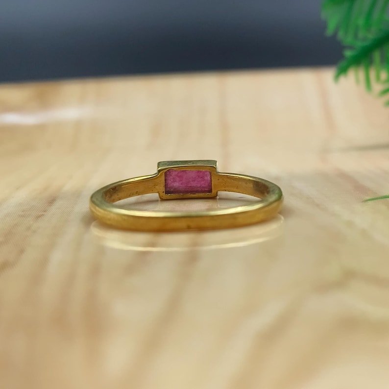Pu&ograve; includere: Un anello d'oro con una gemma rosa rettangolare. L'anello ha una fascia semplice e un'incastonatura a castone per la gemma. L'anello &egrave; mostrato su una superficie di legno di colore chiaro.
