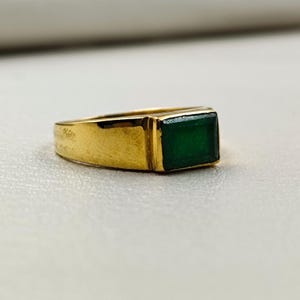 May include: A gold ring with a rectangular, emerald-green gemstone. The ring has a simple, classic design with a polished gold band. The gemstone is set flush with the band, creating a sleek, modern look. The ring is likely a piece of jewelry.